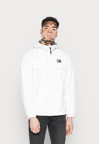 Mladý muž s vitiligem, oblečený v bílé kapucové bundě značky Tommy Jeans a černých džínách, stojící před jednobarevným světlým pozadím.