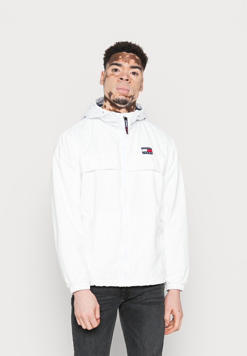 Jonge man met vitiligo in een witte Tommy Jeans capuchonjack en zwarte jeans, staand tegen een effen lichte achtergrond.