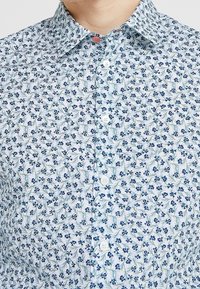 Camicia bianca con bottoni caratterizzata da un motivo floreale blu con steli verdi. Il materiale sembra liscio, con un colletto classico e chiusura con bottoni standard.