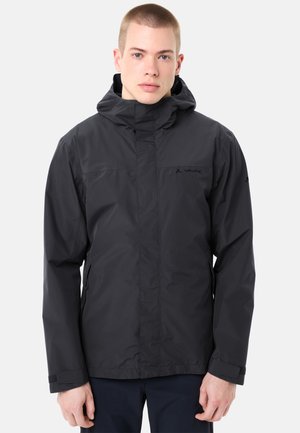 Junger Mann trägt eine schwarze, wasserdichte Kapuzenjacke und steht vor einem einfarbigen, hellen Hintergrund.