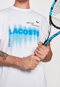 Vit t-shirt med blå gradient "LACOSTE" text, och "SEE YOU ON COURT" ovanför. Bärs med ett svart och blått tennisracket.