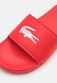 Chancla roja con una tira ancha que presenta un logo de cocodrilo blanco. Hecha de material sintético suave con una plantilla texturizada.