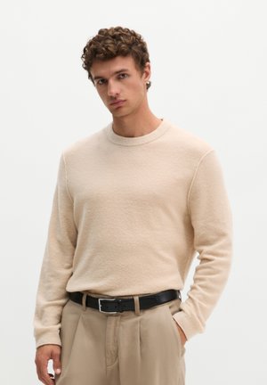 Jeune homme aux cheveux bouclés portant un pull beige rentré dans un pantalon beige avec une ceinture noire, debout devant un fond clair uni.