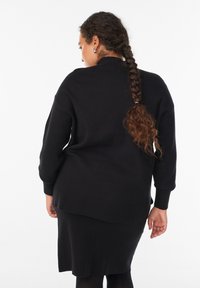 Kobieta z długimi warkoczami, ubrana w czarny sweter i spódnicę, odwrócona plecami na tle jednolitego białego tła.