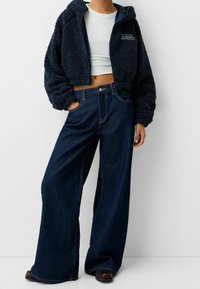 Avkortad navy faux fur-hoodie med huva och broderade detaljer, kombinerad med vida mörk denimjeans med fickor och synlig söm.