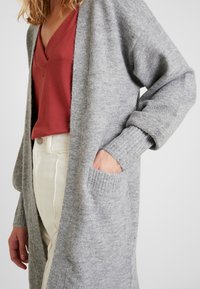 Grå stickad kofta med öppen front, långa ärmar, ribbade ärmslut och sidofickor. Bärs över en röd topp och ljusa byxor.