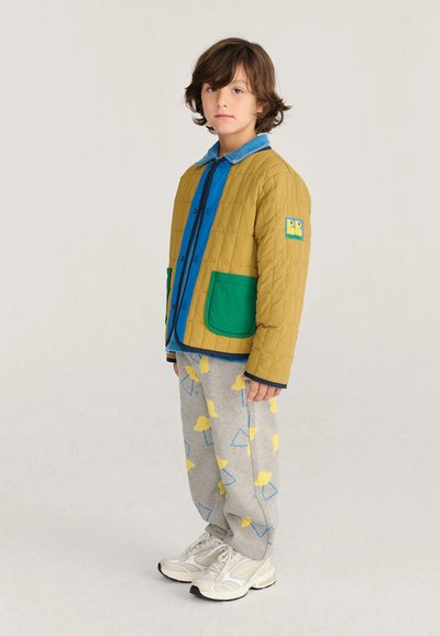 Enfant aux cheveux bruns portant une veste moutarde avec des poches vertes, une chemise bleue, un pantalon gris avec des imprimés d'oiseaux jaunes, et des baskets blanches, debout de profil.