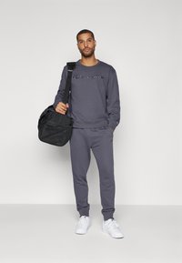 Siv sweatshirt in trenerke z logotipom, v kombinaciji z belimi supergami. Model drži črno torbo za trening z nastavljivim trakom.