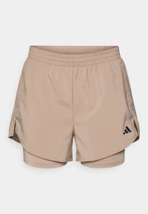 Pantaloni scurți atletici beige cu un brâu, având slits laterale și pantaloni scurți interni. Logo-ul Adidas negru pe partea stângă jos. Textură de material neted.