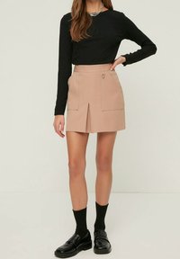 Mujer con una falda mini beige con abertura frontal y bolsillos, top negro de manga larga, calcetines negros y mocasines negros, de pie con una mano en la cadera.