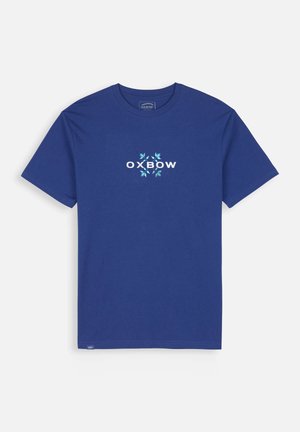 T-shirt en coton bleu à manches courtes et col rond. Avec le texte blanc "OXBOW" et des motifs de feuilles dans des nuances de bleu et de vert.