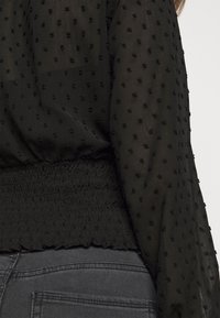 Blusa nera in tessuto testurizzato con accenti a forma di punto. Dettaglio in vita arricciata, maniche lunghe con un aspetto trasparente, abbinata a jeans scuri.