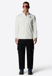 Chaqueta de forro polar blanca con cremallera y bolsillos laterales, combinada con pantalones cargo negros que tienen grandes bolsillos. El modelo lleva gafas de sol y zapatos azules.