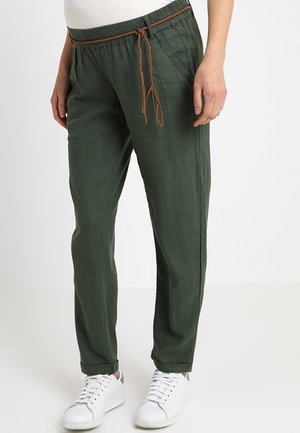 Osoba ubrana w ciemnozielone spodnie ze sznurkiem w pasie z brązowym plecionym wiązaniem, w białych trampkach, stojąca na jasnym, jednolitym tle.