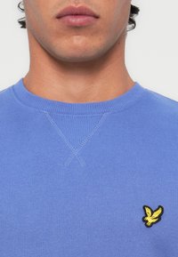 Lyseblå genser med rund hals laget av en myk stoff, som har en gul brodert logo på venstre bryst og ribbestrikkede detaljer på kragen.