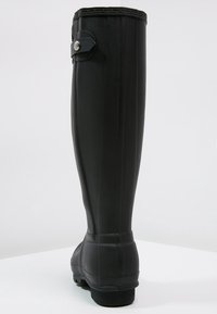 Bota de goma negra hasta la rodilla con una textura suave, punta redondeada, tira decorativa con un botón a un lado y un ligero tacón.