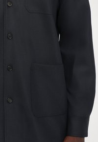 Zblízka na tmavomodrý saká s gombíkmi, ktoré má veľkú prednú kapsu a dlhé rukávy na osobe s tmavou pokožkou.