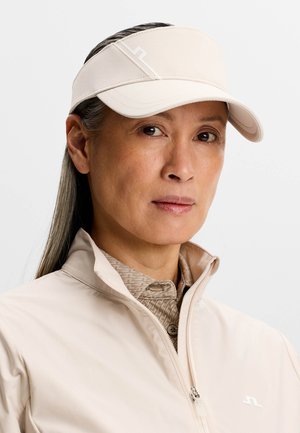 Femme aux longs cheveux gris portant une visière beige, une veste beige zippée et une chemise à col beige à motifs, sur un fond uni.