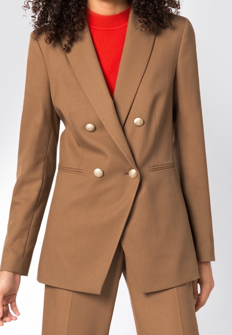 Brun dubbelknäppt kavaj med guldknappar, strukturerad design, något texturerad tyg och slagkrage, bärs över en röd topp.