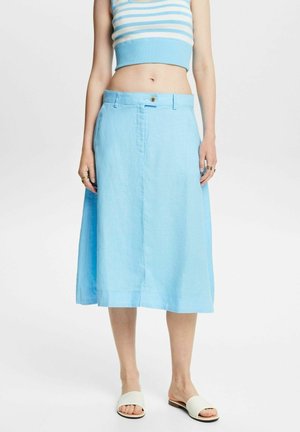 Femme portant une jupe midi bleu clair, un débardeur court rayé bleu et blanc, des sandales blanches et des bagues en or aux doigts.