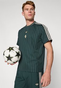 Mørkegrøn stribet kortærmet skjorte med en sort-hvid fodbold. Funktionen inkluderer to cremefarvede striber på skuldrene og broderede detaljer.