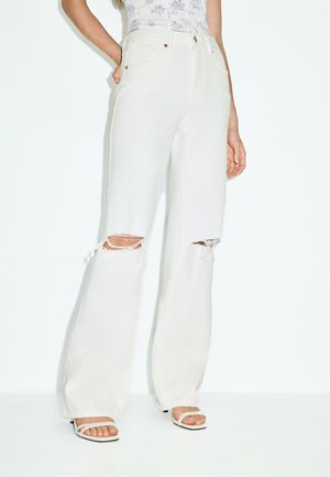 Femme portant un jean blanc taille haute à jambes larges avec des genoux déchirés et des sandales blanches à bouts ouverts, debout devant un fond uni.