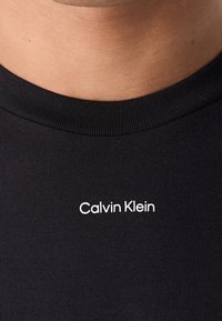 T-shirt en coton noir avec col rond. Logo blanc "Calvin Klein" centré sur la poitrine. Texture lisse.