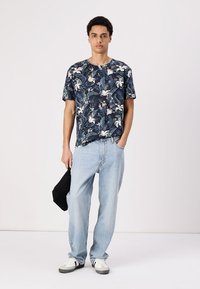 Cămașă florală cu mânecă scurtă, de culoare albastru închis, cu model verde și alb, asociată cu blugi ușor albaștri și adidași albi, purtând o geantă neagră.