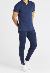 Mies seisoo kädet selän takana, päällään tummansininen polo-paita, vastaavat sivuinsigneeratut jogger-housut, valkoiset sukat ja valkoiset lenkkarit.