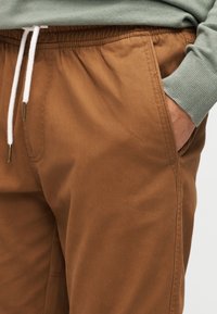 Καφέ βαμβακερό παντελόνι jogging με ελαστική ζώνη μέσης, λευκό κορδόνι, πλαϊνή τσέπη και ορατές ραφές. Μαλακή υφή και άνετη, καθημερινή εφαρμογή.