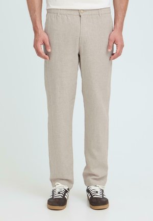 Mężczyzna ubrany w beżowe proste spodnie i czarne trampki z białymi sznurówkami, stojący z rękami swobodnie opuszczonymi wzdłuż ciała.