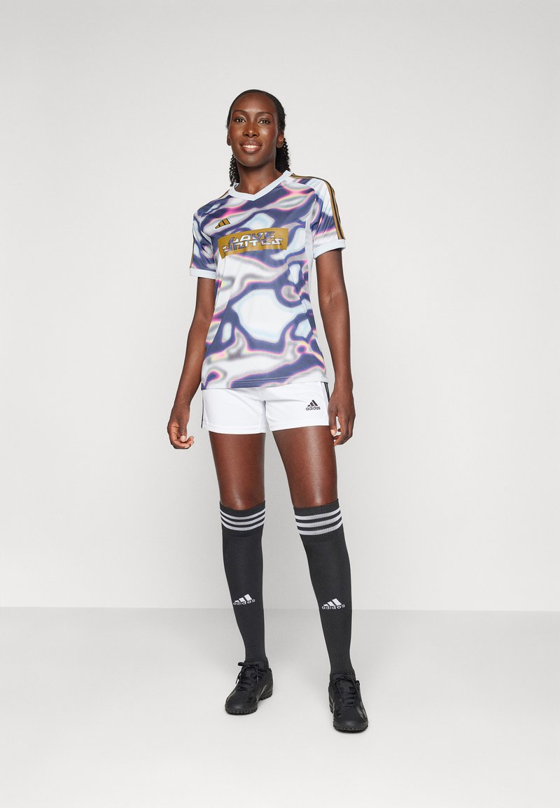 Frau in einem mehrfarbigen Fußballtrikot mit dem Aufdruck "Love Unites", weißen Shorts, schwarzen Adidas-Kniestrümpfen und schwarzen Fußballschuhen, stehend vor weißem Hintergrund.
