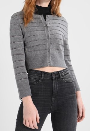 Femme portant un cardigan court gris boutonné avec des rayures texturées horizontales sur un haut noir, assorti à un jean gris foncé taille haute.