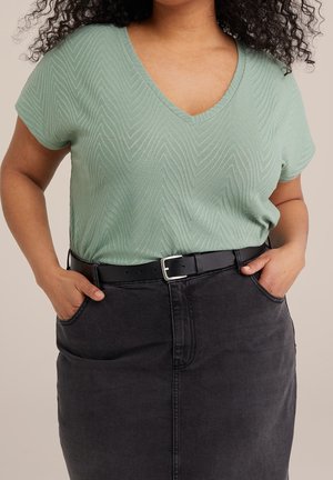 Femme portant un haut vert clair texturé à col en V, rentré dans une jupe en jean noire avec une ceinture noire, mains dans les poches de la jupe.