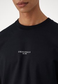 Fekete pamut póló, kerek nyakkal. A közepén fehér nyomott szöveg látható: "ORIGINALS est. 1990". Sima tapintású.