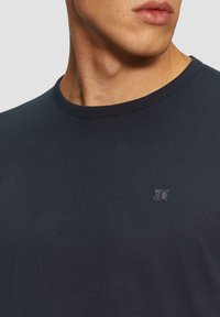 Navyblaue Baumwoll-T-Shirt mit Rundhalsausschnitt und kurzen Ärmeln, mit einem kleinen geprägten Logo auf der Brust und einer glatten, gleichmäßigen Textur.