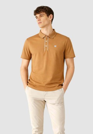 Giovane uomo con polo marrone a maniche corte con bottoni bianchi e pantaloni beige, in piedi con le mani nelle tasche davanti a uno sfondo grigio.