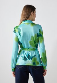 Bluza florală de un albastru deschis, dintr-un material neted, cu modele de flori verzi, guler, mâneci lungi și talie strânsă cu o panglică.
