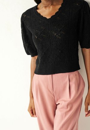 Top de punto negro de manga corta con patrón texturizado y escote con ondas, combinado con pantalones de talle alto rosas claros con pliegues.