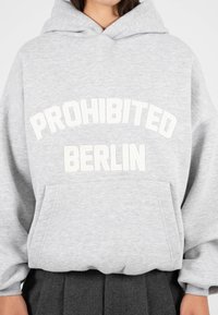 Šedá mikina s kapucí a přední kapsou, na které je bíle vyšitý nápis "PROHIBITED BERLIN." Obsahuje stahovací kapuci a žebrované manžety.