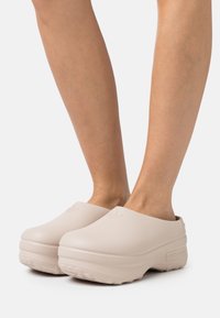 Světle růžové dřeváky s hladkým, zaobleným designem a texturovanou podrážkou. Mají vyražené logo a mírně zvýšený podpatek.