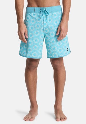 Mężczyzna ubrany w niebieskie kąpielówki z białym kwiatowym wzorem, stojący boso na białym tle.