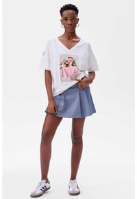 Witte, oversized t-shirt met een kleurrijke grafische print op de voorkant, gecombineerd met een blauwe geplooide rok en witte sneakers met zwarte strepen.