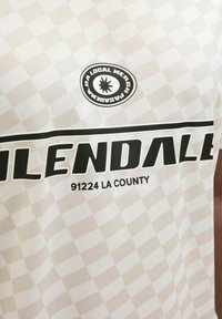 Valge ruuduline T-särk, millel on julged mustad kirjutas "GLENDALE 91224 LA COUNTY" ja ümmargune logo, millel on päikesepaiste disain.