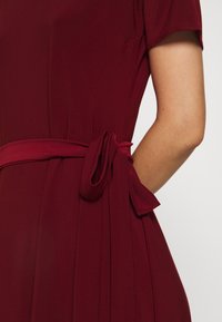 Bordeauxklänning med korta ärmar, som har en plisserad design och en knytberlock i midjan, gjord av ett mjukt och smidigt tyg.