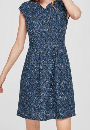 Vrouw draagt een knielange blauwe bloemenjurk met cap sleeves en een getailleerde taille, staand tegen een effen achtergrond.