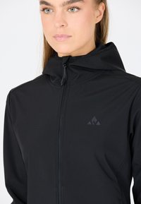 Veste noire à capuche avec une texture lisse et mate.Elle présente une fermeture éclair à l'avant, un logo subtil sur la poitrine et un design ajusté.