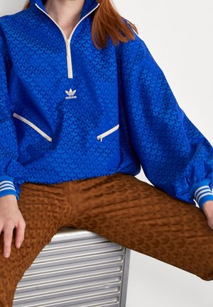 Persona che indossa una giacca Adidas blu con cerniere bianche a metà, e pantaloni marroni a fantasia, seduta su una superficie metallica.
