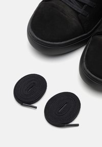 Svarta mockaskor med två spiralformade svarta skosnören placerade i närheten på en vit yta.