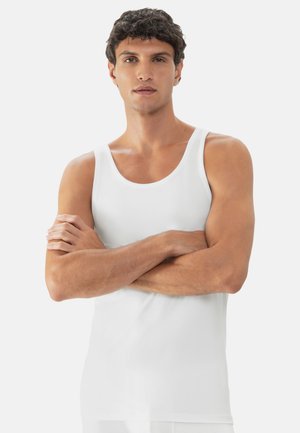 Ung mand med kort, mørkt krøllet hår iført en hvid tanktop, stående med armene krydset foran en ensfarvet hvid baggrund.